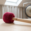 Basic Mallet mit weinrotem Fleecekopf und Holzgriff, im Hintergrund ein Gong im hellen Raum